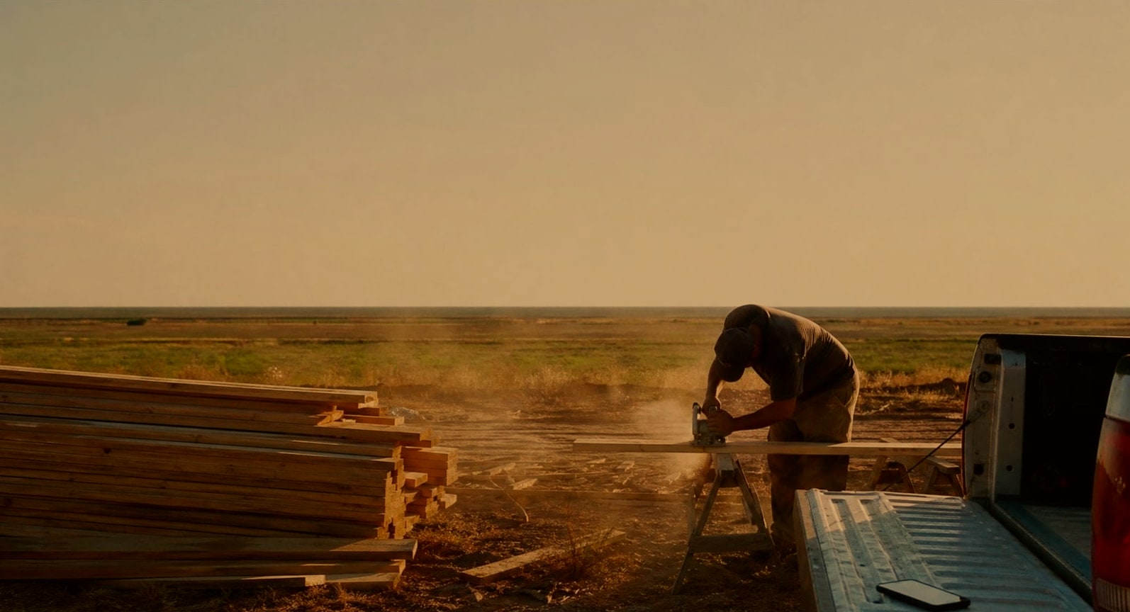 A framer bent over a sawhorse cutting a board on a flat, hazy job site in low afternoon sun, lumber stacked beside him and a phone lying on a truck tailgate out of reach.